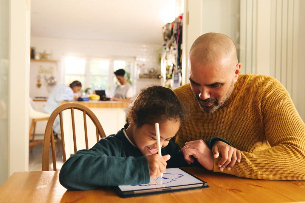 Photo of a child drawing on an ipad at the table sitting next to their Dad