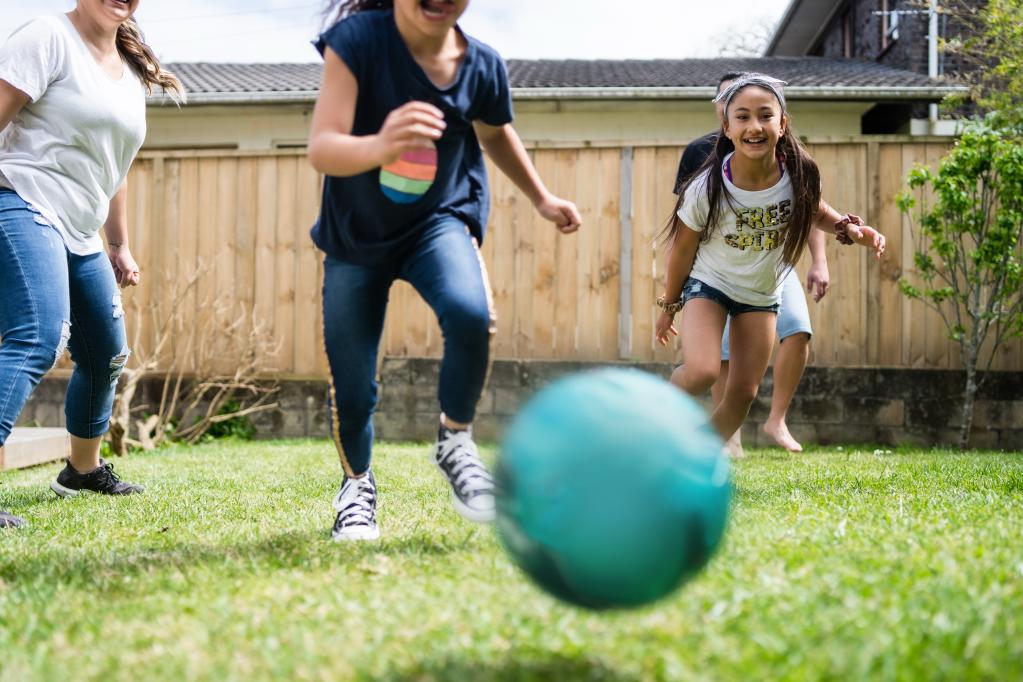 A group of children are playing with a blue ball on the grass in a backyard. They are smiling and running toward the camera, enjoying an active game together on a sunny day.