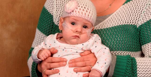 This image shows a baby wearing a white crocheted hat with a pink flower, sitting in the arms of an adult. The baby has nasal oxygen tubes and appears alert, with a calm expression, while the adult is partially visible wearing a green-and-white striped knit sweater.