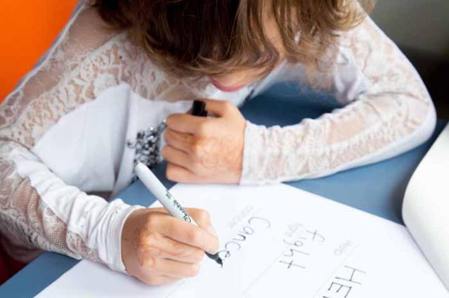 A child with wavy brown hair is sitting at a table, writing in black marker on a sheet of paper that includes the word "Cancer." The child is focused on their work, wearing a long-sleeved white shirt with lace details.