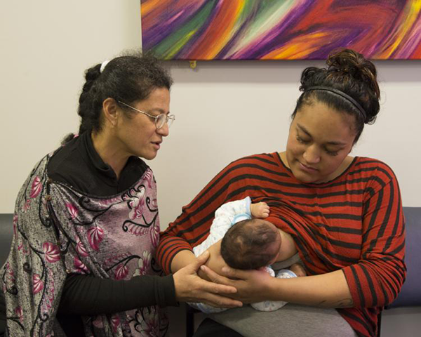 Mother breastfeeding her baby, in rugby hold. Lactation consultant is looking on. 