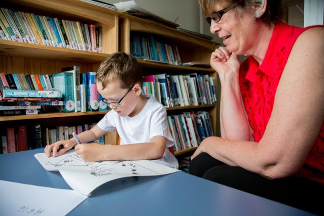 A young boy with glasses is seated at a table in a library, focused on drawing in a book, while an older woman in a red shirt watches and smiles beside him. The background shows shelves filled with books.