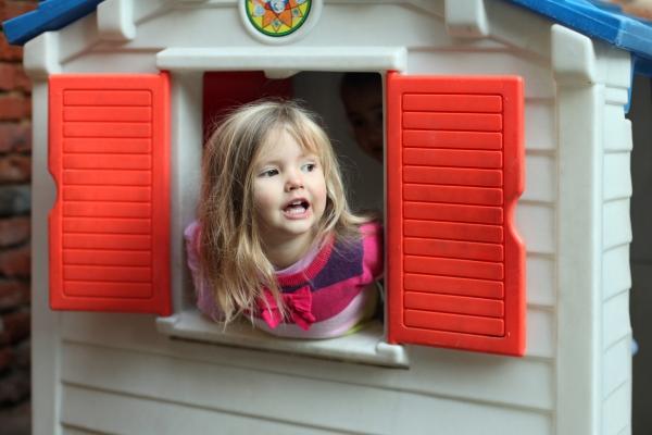 A young girl with long blonde hair is leaning out of a plastic playhouse window with red shutters, smiling.