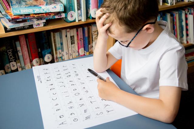 A young boy wearing glasses sits at a table in a library, looking frustrated as he works on a large worksheet with alphabet letters. He is holding a pen and has one hand on his head, appearing to struggle with the task.