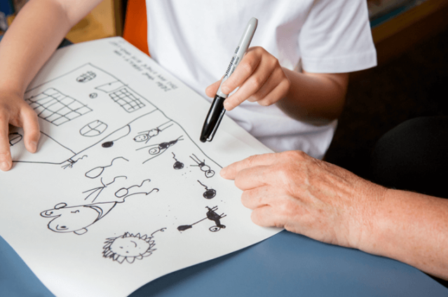 A child is holding a black marker and showing a drawing to an adult. The drawing depicts a house and several stick figures, animals, and objects. The adult is pointing at part of the drawing.