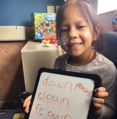 A young girl with a nasal tube is sitting and smiling while holding up a small whiteboard. The whiteboard displays the words "down," "clown," and "fr_own" written in orange marker. A box of crayons and art supplies is visible in the background.