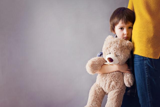 Little brown haired boy, hugging his mother's leg and a teddy bear with a worried look on his face.