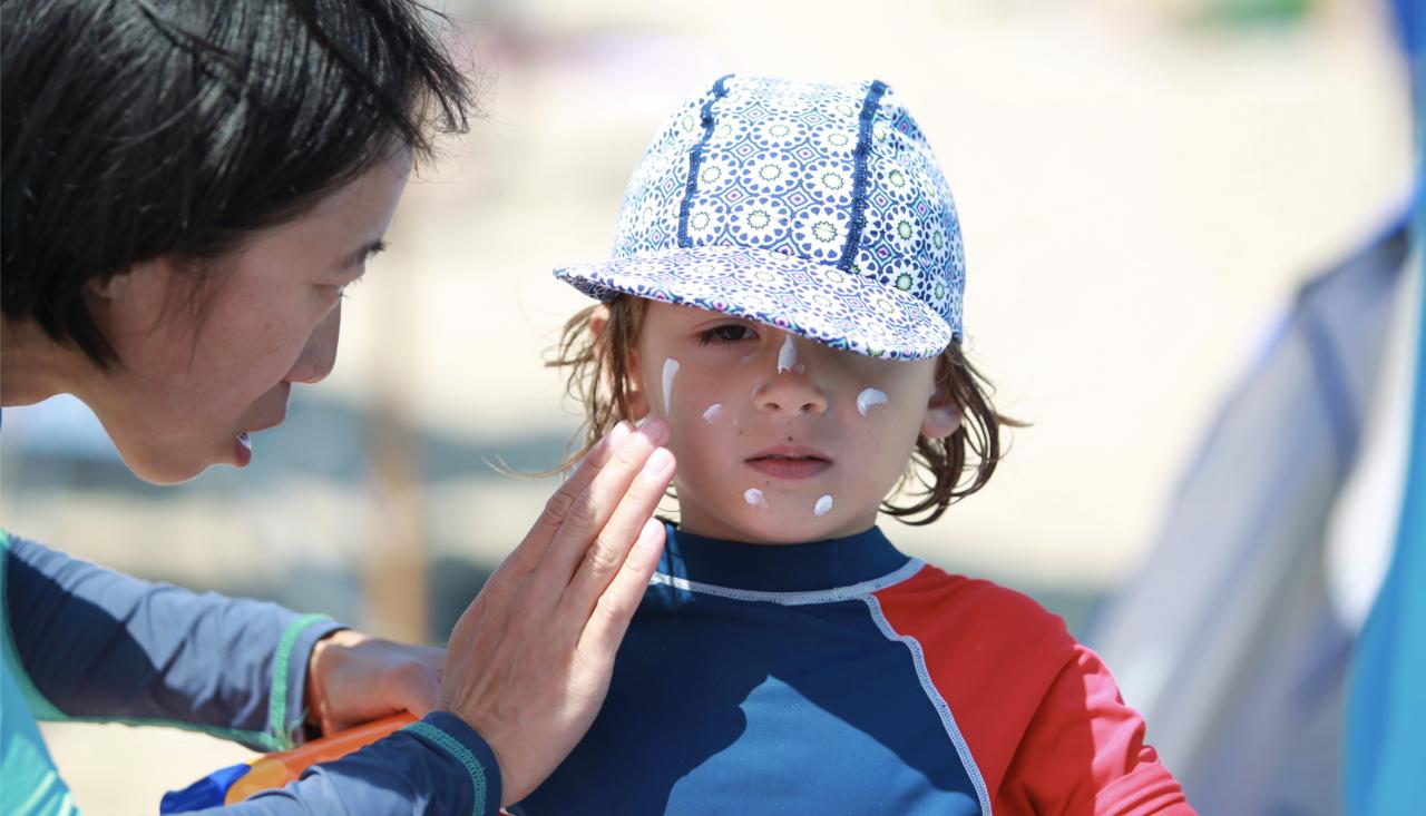 Image of a child in a hat and rash vest having sunblock applied to their face by a parent